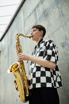 Vertical shot of young adult Caucasian man playing saxophone outdoors near concrete wall. Image suitable for music education marketing, street performance promotion, or cultural event advertising