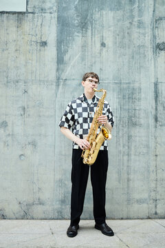 Vertical shot of young Caucasian man playing saxophone against concrete wall, representing street musicians and urban culture. Image is ideal for music festival promotion