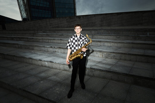Portrait of Caucasian teenager holding saxophone on city stairs, performing as street musician. Image suitable for youth music education and urban lifestyle marketing, high angle shot