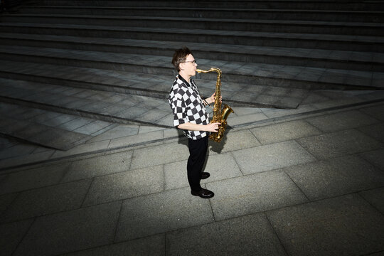 Young adult Caucasian man playing saxophone on city stairs, performing as street musician. Image suitable for music festival promotion or urban lifestyle marketing, high angle shot