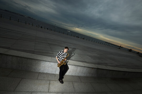 Young adult Caucasian man playing saxophone on urban waterfront promenade under dramatic sky. Image suitable for music festival promotion or creative arts marketing, high angle shot