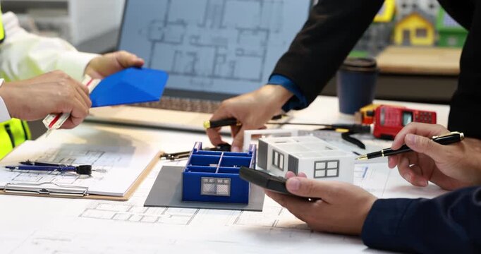 Architects and construction workers review house plans with a miniature model, blueprints, tools and paperwork on a busy office desk.