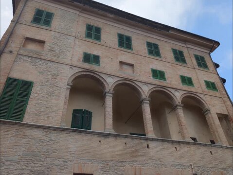 Facciata di un palazzo storico in mattoni con loggiato ad archi, colonne classiche e persiane verdi sotto un cielo parzialmente nuvoloso.