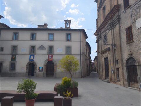 Piazza storica di un borgo italiano con palazzi antichi, un piccolo campanile a vela e vasi decorativi sotto un cielo luminoso.