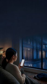大雨の夜に窓外を確認しながらスマートフォンを持つ女性