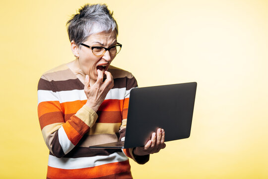 Elderly woman with short grey hair and glasses expressing shock and frustration while looking at a laptop computer