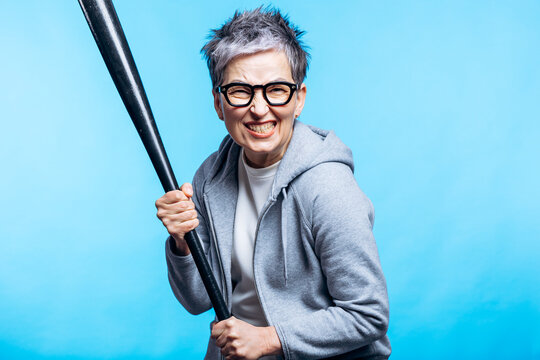Senior woman wearing glasses and hoodie, clenching teeth and gripping a baseball bat, expressing anger on blue background