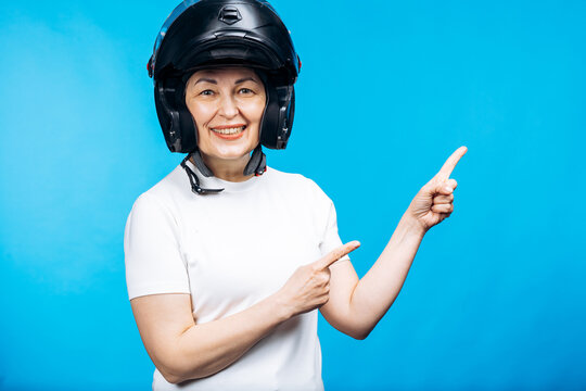 Mature woman wearing a black motorcycle helmet and white t-shirt, smiling and pointing at blue background with both hands