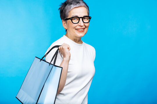 Smiling senior woman with eyeglasses holding a blue shopping bag, enjoying retail therapy and consumerism