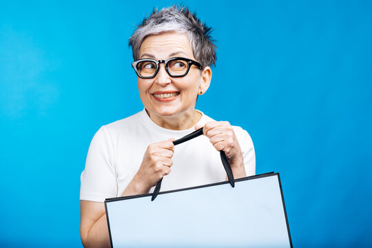 Happy senior woman with gray hair wearing glasses holding a shopping bag, looking to the side with excitement on blue background