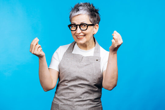 Smiling senior woman wearing apron and glasses making an expressive italian hand gesture, standing on a blue background