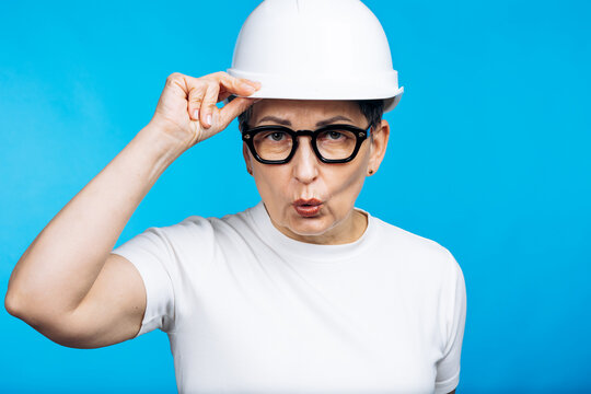 Experienced woman engineer wearing a white hard hat and glasses, posing with an expressive face against a bright blue background