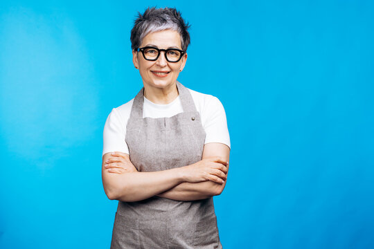 Senior woman wearing an apron and eyeglasses, standing confidently with arms crossed against a vibrant blue background, looking at the viewer