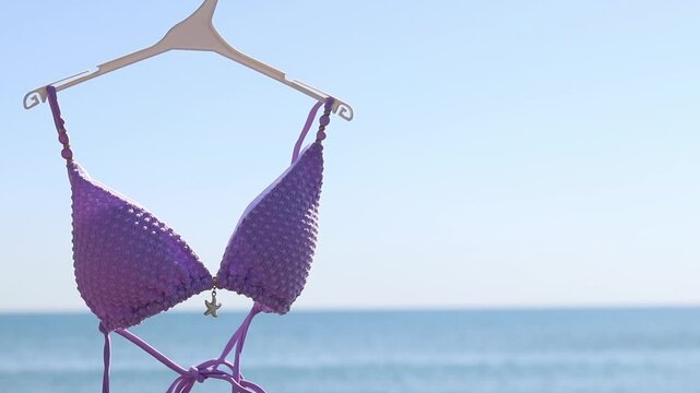 Lilac swimsuit-bodice on a hanger against the background of the blurred sea. Top from a women's separate swimsuit of a classic curtain shape in lavender color. Summer vacation concept