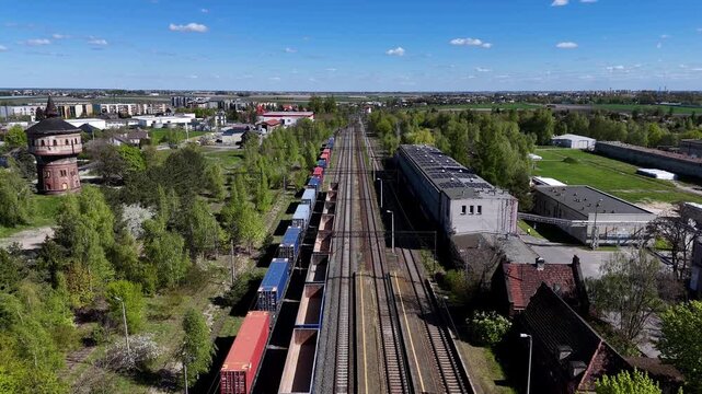 Stationary freight train with colorful cargo containers and open hopper cars parked on railway station tracks near historic brick water tower, industrial buildings, and green trees.