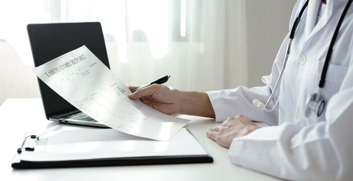 Doctor holding laboratory report document and pen to analyze medical patient health results near laptop on desk in hospital clinic office space