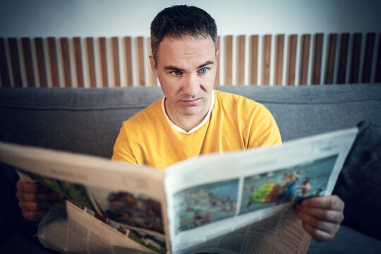 Surprised man reading a newspaper at home, reacting to breaking news while sitting on sofa in a modern living room.