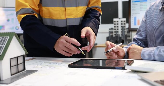 Engineers review building plans with a tablet and model house in an office, discussing a residential construction project.