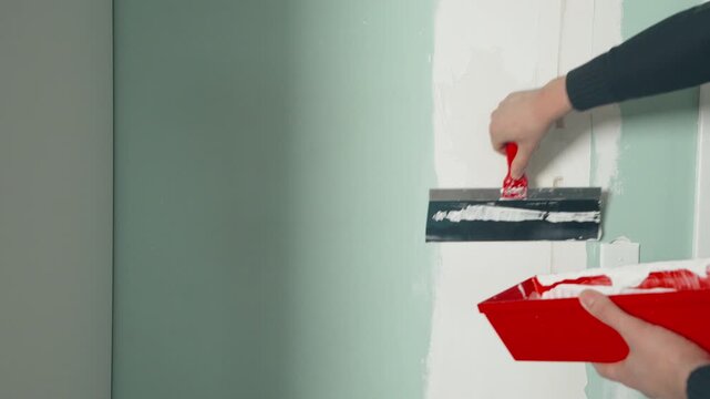 Hands loading drywall compound onto a wide putty knife from a red mud pan near a freshly patched wall
