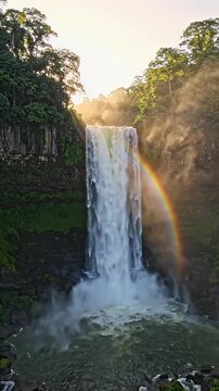 Majestic Waterfall at Sunrise with Rainbow - A breathtaking waterfall cascades down a cliff face, illuminated by the warm glow of a rising sun.
