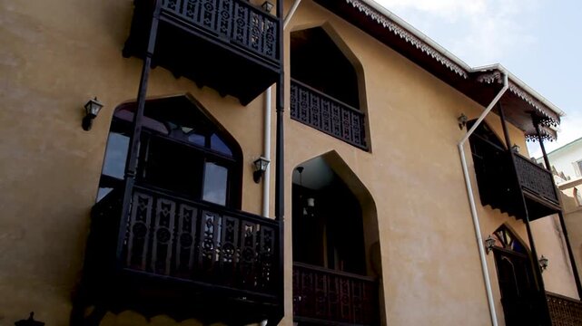 A low-angle shot of a traditional building featuring warm-toned stucco walls, intricate dark wooden balconies, and distinct pointed arched windows.