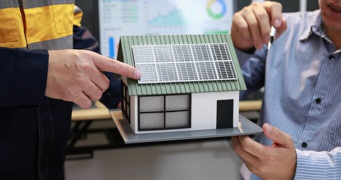 Engineers discuss a small solar-powered house model in an office, pointing at roof panels while reviewing energy data on a computer screen.