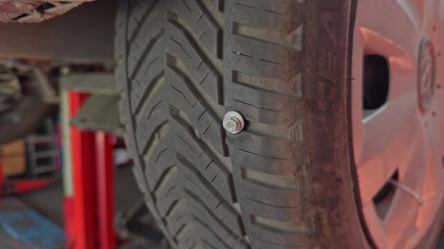 Close up view reveals a metal bolt embedded in the tread of a car tire on a lift in an auto repair garage