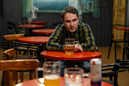 Man sitting alone at a bar with a mug of beer, looking at the camera with a tired, lonely expression. Solitary drinking, loneliness, and alcohol habit. man drinks beer in a bar
