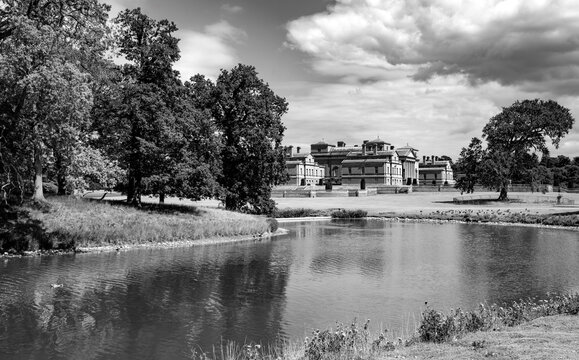 Black and white view of Holkham Hall across the lake in Holkham Park, Norfolk, England. The Palladian country house is surrounded by historic parkland and mature trees under dramatic clouds.