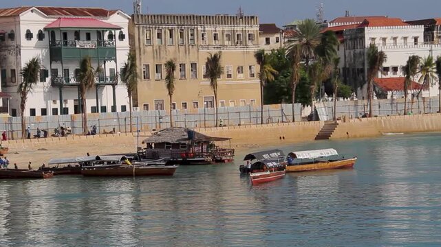 A scenic view of traditional wooden dhow boats docked along the sandy shore of Stone Town, Zanzibar, with historic colonial architecture in the background.