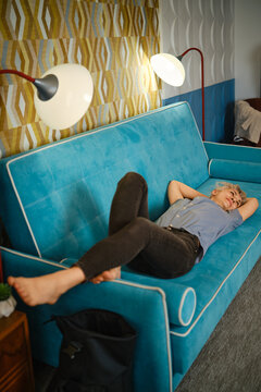 Person rests on blue couch with bare feet up, arms behind head. Room has warm light from lamps and decorative wall behind couch. Scene captures casual and comfortable afternoon.