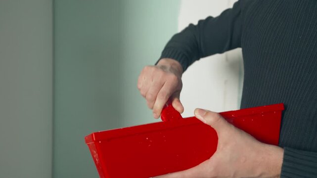 Man in dark sweater holding a putty knife and red drywall mud pan preparing to patch a wall