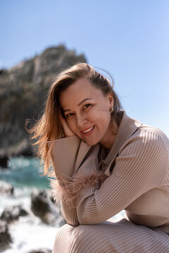 Smiling mature woman posing gracefully in beige ribbed outfit near rocky seaside coastline on sunny day