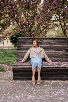 Young woman resting on wooden bench beneath blooming cherry tree branches in city park during pleasant spring weather