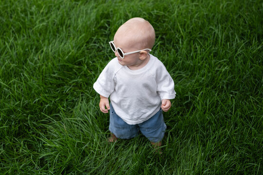 A charming boy wearing sunglasses is learning to walk on the green grass. The little one is walking barefoot on the lawn.