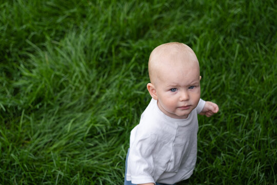 A charming boy wearing sunglasses is learning to walk on the green grass. The little one is walking barefoot on the lawn.