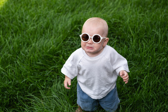 A charming boy wearing sunglasses is learning to walk on the green grass. The little one is walking barefoot on the lawn.