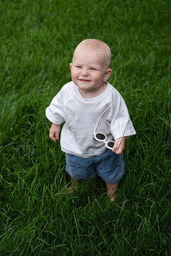 A charming boy wearing sunglasses is learning to walk on the green grass. The little one is walking barefoot on the lawn.