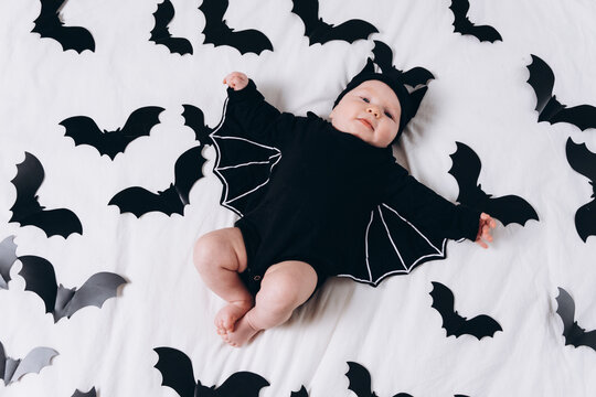 A cute baby in a bat costume lies on a white background featuring a bat pattern. A Halloween costume for a baby.