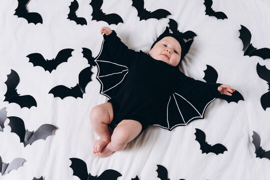 A cute baby in a bat costume lies on a white background featuring a bat pattern. A Halloween costume for a baby.