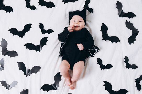 A cute baby in a bat costume lies on a white background featuring a bat pattern. A Halloween costume for a baby.
