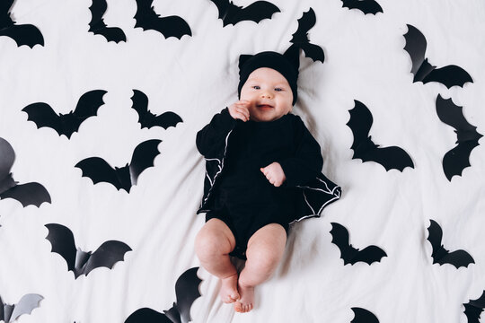 A cute baby in a bat costume lies on a white background featuring a bat pattern. A Halloween costume for a baby.