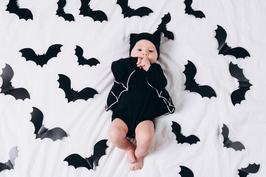 A cute baby in a bat costume lies on a white background featuring a bat pattern. A Halloween costume for a baby.
