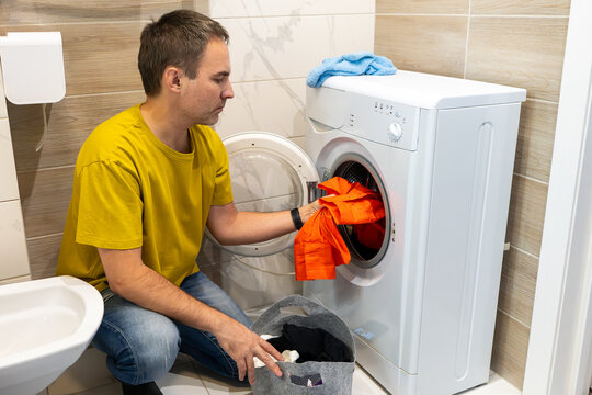 A man in a yellow t shirt loads an orange garment into a washing machine; a blue towel rests on top, a laundry basket stands nearby, in a compact tiled bathroom.