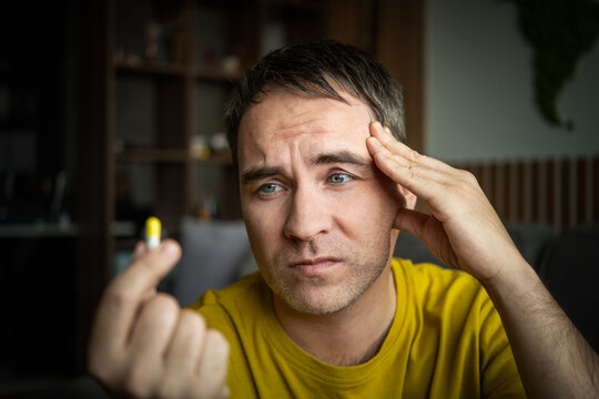 Man suffering from a severe headache, holding his head with one hand and showing a yellow pill. Migraine, stress, and medication for pain relief.