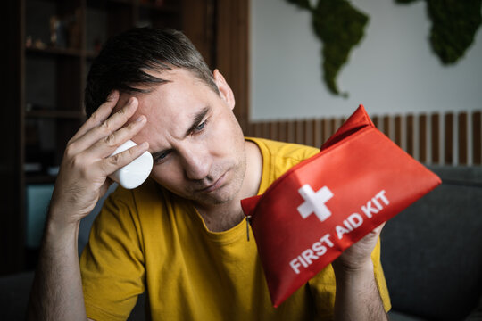 Man searching for painkillers in a red first aid kit, suffering from a severe headache. Migraine, head pain, and urgent medical treatment.