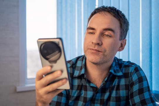 Man checking smartphone screen indoors with thoughtful expression near window blinds in casual plaid shirt, with man, smartphone, phone, looking detail, showing checking, screen, indoors, window