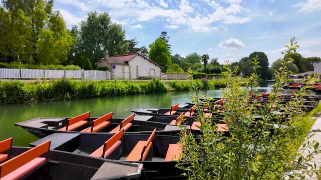 Rows of small black boats with orange seats are moored along a calm river beside village houses, trees and gardens, creating a detailed waterside scene under a bright blue sky