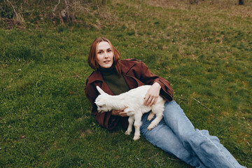 Naklejka premium Relaxed woman lies on grass with a playful goat kid beside her, capturing countryside vibes and gentle companionship. Casual fashion, cozy mood, and natural lighting convey a wholesome lifestyle