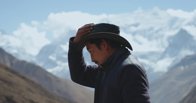 Annapurna Circuit, Manang, Nepal, camera shot of a local Sherpa man putting on and adjusting a cowboy-style hat, against a blurred background of towering Annapurna peaks and snow-dusted Himalayas